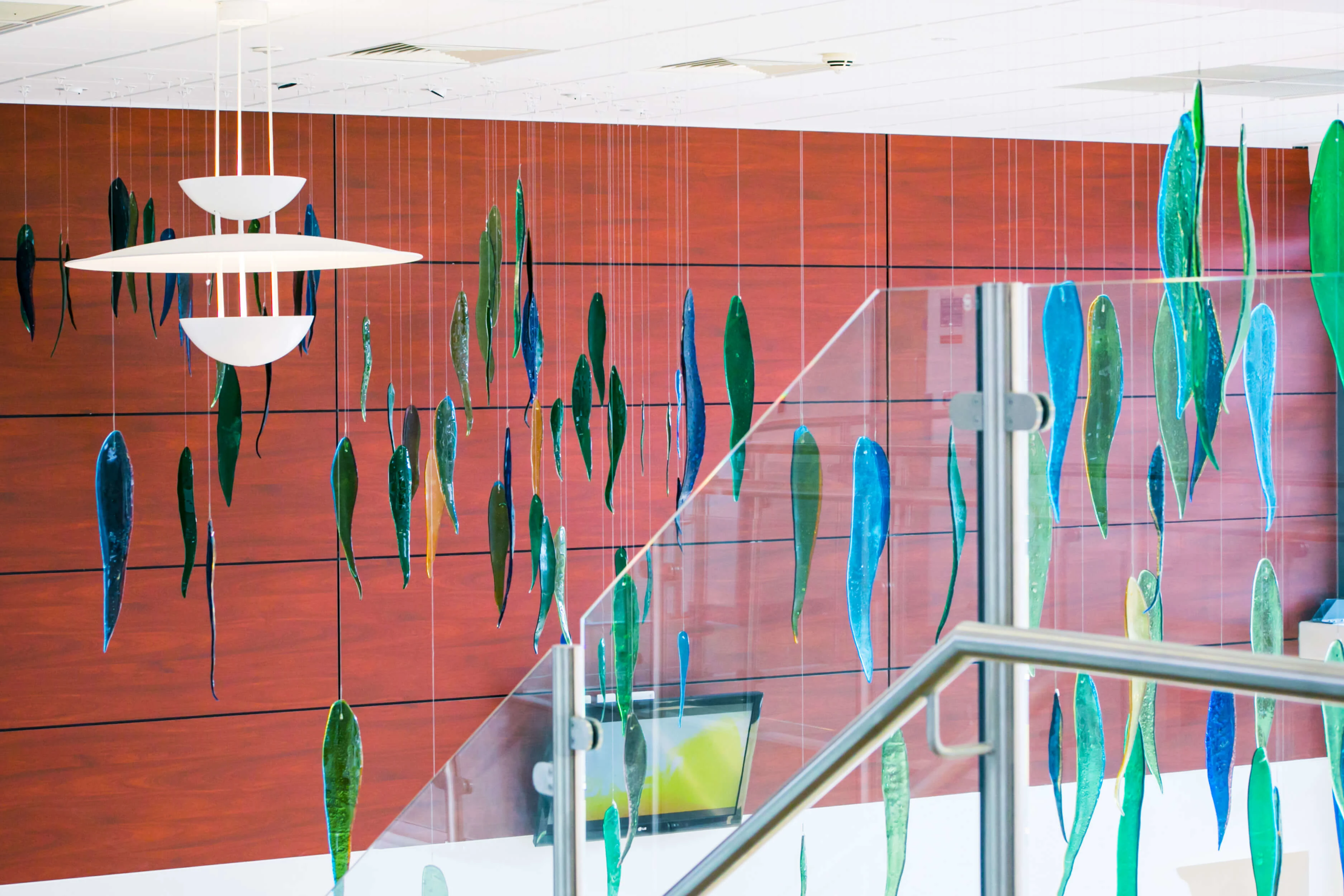 Artwork with a school of fish hanging from a ceiling. The wall behind it has a warm auburn colour. The fish are a mixture of colours - either blue or green. The depth of colour changes depending on the light.