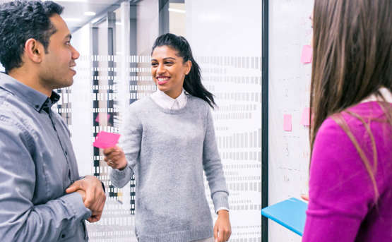 3 colleagues in an office environment working on a collaborative task. One of the colleagues is smiling at her colleague while's she's handing him a pink post-it