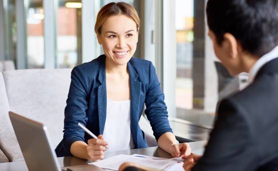 a smiling woman signs paper in front of a man wearing a suit.