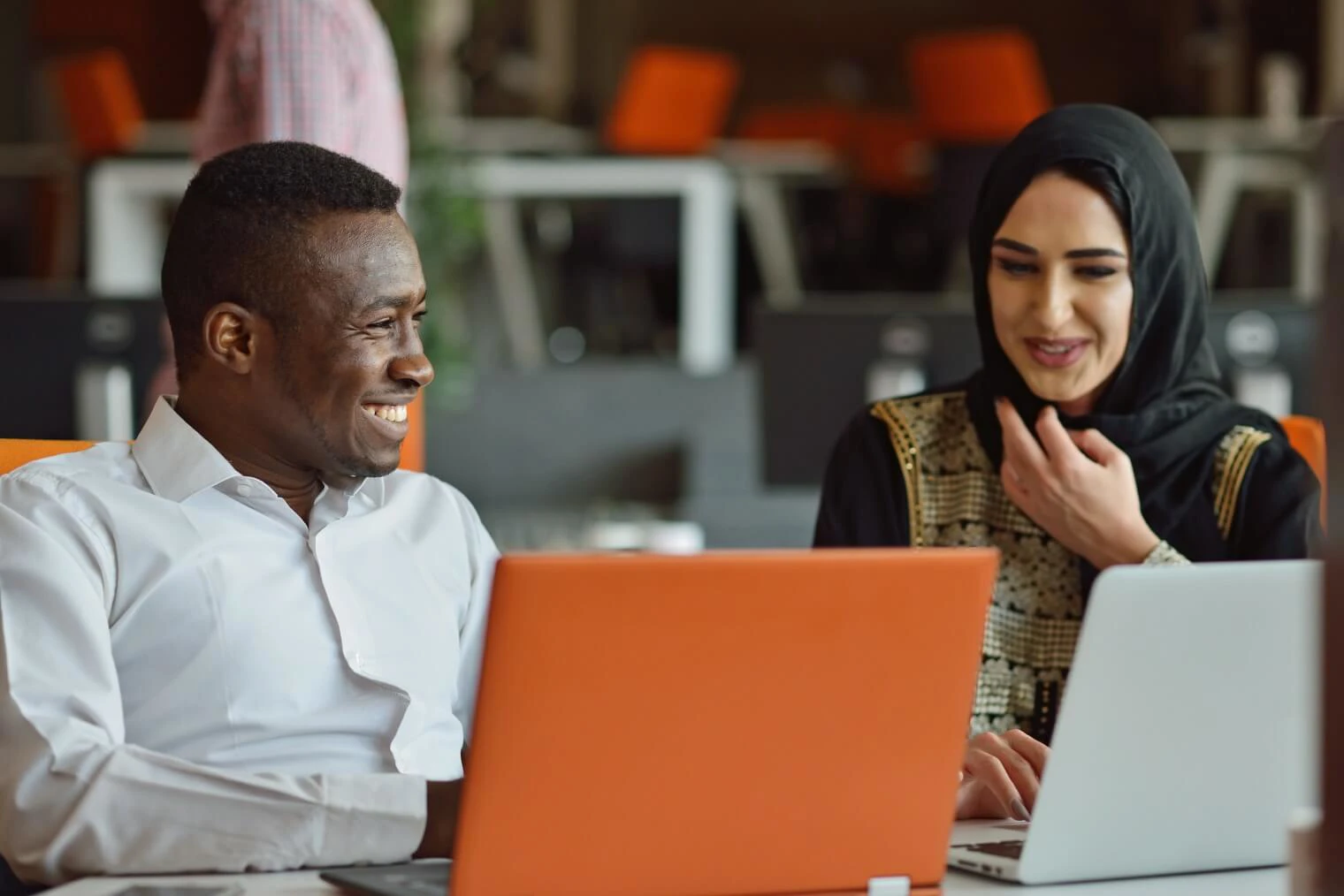 a woman wearing a hijab smiles at a colleague as they work from laptops 
