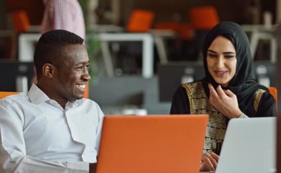 a woman wearing a hijab smiles at a colleague as they work from laptops 
